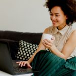 A woman sitting on a couch using a laptop while drinking a coffee